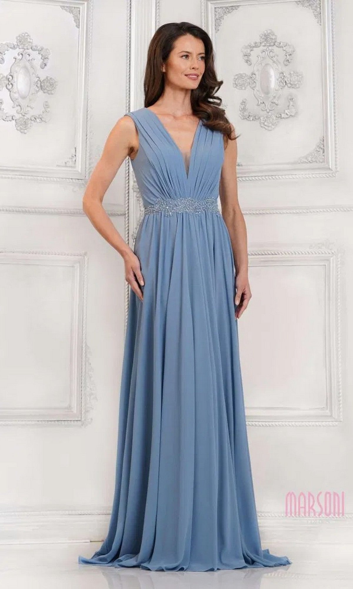 A woman in the Marsoni MV1292 long formal dress, featuring a sleeveless, floor-length light blue chiffon design with V-neckline and ruched bodice, stands against an ornate white wall.